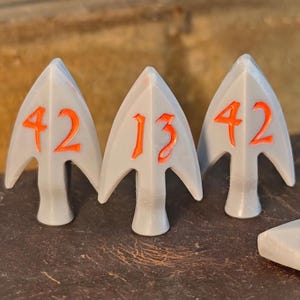 Puede incluir: Tres puntas de flecha grises dispuestas verticalmente, cada una con un número grabado en naranja brillante. Las puntas de la izquierda y la derecha están marcadas con el número 42, mientras que la central muestra el número 13 en una fuente estilizada.