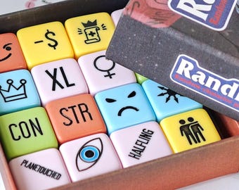 Puede incluir: Un conjunto de dados cuadrados coloridos dentro de una caja oscura etiquetada como Randice. Los dados presentan varios símbolos y textos impresos en negro, incluyendo XL, CON, STR, HALFLING, PLANETOUCHED, una corona, un ojo y expresiones.