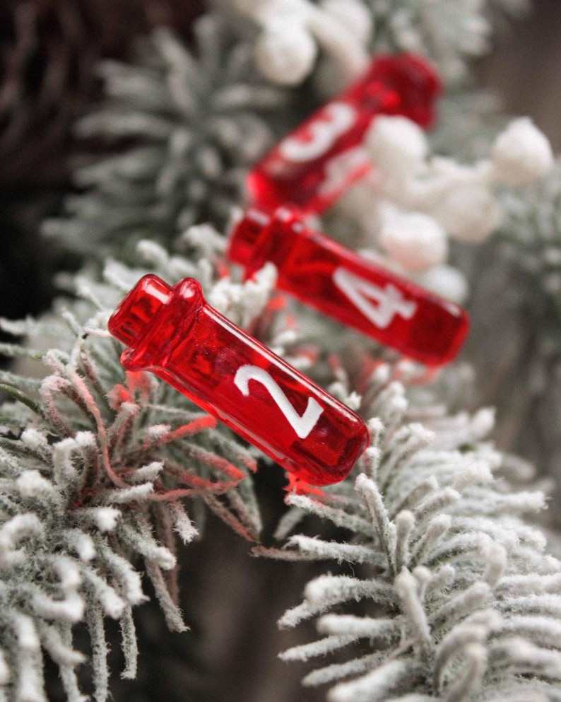 A close-up photo of red translucent vial-shaped D4 dice nestled among frosted, snow-dusted evergreen branches. The nearest vial shows a bold white number 2, with two more vials showing 3 and 4 blurred in the background. A dark pinecone and white berries are also visible in the softly blurred background, giving a festive winter holiday feel.