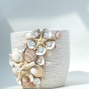 May include: White ceramic planter with a rope-like texture. The planter is decorated with seashells and faux pearls. The seashells are arranged in a cluster around a starfish.