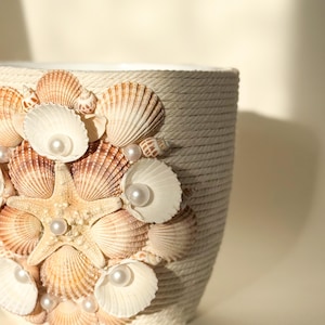 May include: A white ceramic planter decorated with seashells and faux pearls. The planter has a rope-like texture and is a great addition to a coastal-themed home decor.