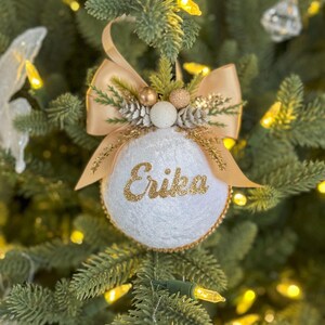 May include: White felt Christmas bauble with a gold glitter bow and the name "Erika" written in gold glitter. The bauble is decorated with gold and white beads and pine sprigs.