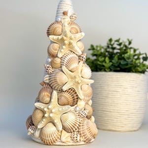 May include: A white cone-shaped Christmas tree decoration covered in seashells and starfish. The tree is decorated with white pearls and has a rope base.