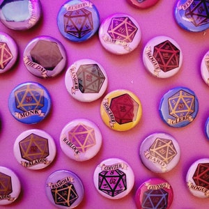 May include: A collection of colorful, round buttons with various Dungeons & Dragons class names and dice designs. The buttons feature text like "Warlock," "Wizard," and "Druid," along with a twenty-sided die graphic. The background is a solid pink.