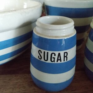 May include: A set of three blue and white striped ceramic kitchen canisters. The canisters are labelled "Sugar", "Tea", and "Coffee".