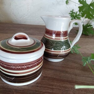 May include: A set of two ceramic kitchenware pieces with a brown, green and white striped design. The set includes a creamer and a sugar bowl with a lid.