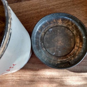 Pu&ograve; includere: Un barattolo di metallo bianco con un'etichetta rossa e un coperchio abbinato. Il coperchio &egrave; leggermente arrugginito e ha una superficie usurata.