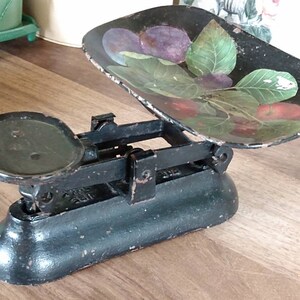 May include: Black cast iron kitchen scale with a painted metal tray featuring a design of green leaves and red and purple fruit. The scale has a round weighing platform and a dial with numbers.