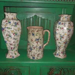 May include: Three vintage ceramic jugs with a floral pattern in shades of blue, green, yellow, and red. The jugs are displayed on a green painted wooden cabinet with carved details.