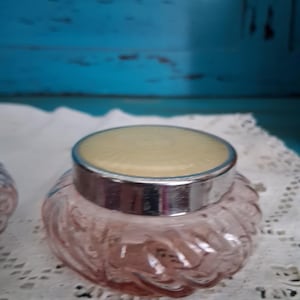 May include: A pink glass jar with a silver lid and a white top. The jar has a ribbed design and is sitting on a white lace doily.