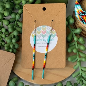 May include: Turquoise, red, yellow, and black beaded earrings hanging from silver hooks. The earrings are displayed on a brown card with the text "beaded by robin" and a website address. The background includes green plants and a wooden surface.