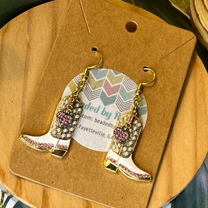 May include: Gold-tone cowboy boot earrings with white enamel, pink and clear rhinestones, and a heart design. The earrings hang from gold-colored hooks. The earrings are displayed on a brown card with text.