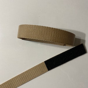 Könnte beinhalten: Ein beigefarbenes Nylonband mit einem schwarzen Klettverschluss. Das Band ist etwa 2,5 cm breit.