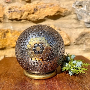 Op de afbeelding: Een ronde, zilveren en gouden bal bedekt met kleine, ronde metalen studs. De bal ligt op een gouden basis en een houten oppervlak.