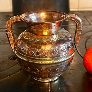 May include: A copper-toned metal vase with two handles and a textured surface. The vase has a bulbous body with ornate detailing and a hammered finish. A bright orange tangerine and a walnut are next to the vase.
