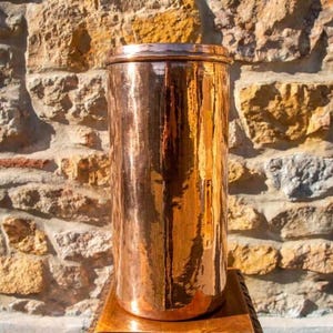 May include: A tall, cylindrical copper container with a polished, reflective surface and a flat, circular lid. The metal body features a smooth, vertical texture with subtle variations in color, ranging from bright orange to deep bronze tones.