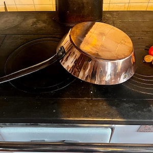 May include: A copper saucepan with a long handle rests on a black stovetop. The shiny pan reflects the surrounding environment. A small tomato, two walnuts, and a green leaf are next to the pan. The stove has a white enamel base.
