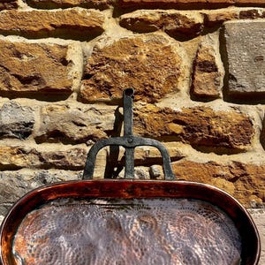 May include: An antique copper and iron oval pan with a decorative handle. The copper pan has a dark, reddish-brown hue with a textured surface. The handle is black and attached to the back of the pan. The background is a rough, textured stone wall.