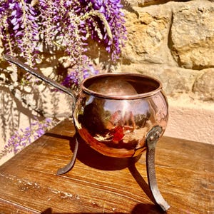 May include: A copper pot with a long handle and three legs. The pot is sitting on a wooden table.