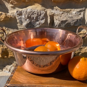 May include: A polished copper bowl with two looped handles, containing several oranges. The bowl is on a wooden surface, with a stone wall behind it. The oranges are a vibrant orange colour.