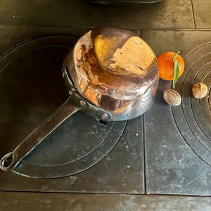 Könnte beinhalten: Eine glänzende Metallpfanne mit langem Griff, die auf einem dunklen Herd steht. Eine Orange und zwei Walnüsse liegen neben der Pfanne. Die Oberfläche der Pfanne reflektiert das Licht.