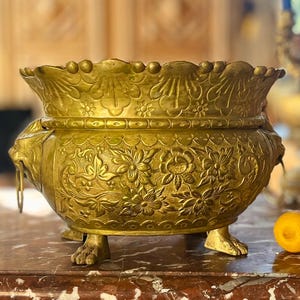 May include: A large, round, gold-colored brass bowl featuring intricate embossed floral patterns and scalloped edges. The bowl is supported by three claw-shaped feet and includes two side handles shaped like lion heads with hanging rings.