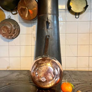 May include: A large, shiny copper pan with a long handle sits on a dark stove top. Other copper cookware hangs on the white-tiled wall behind it. An orange and two walnuts are on the stove top.