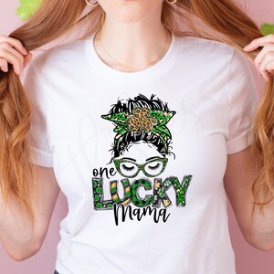 May include: White t-shirt with a green, gold, and black graphic of a woman with a shamrock headband and the text "one lucky mama".