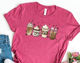 Pode incluir: Uma camiseta rosa com um gráfico de quatro xícaras de café com pegadas de patas e o texto "Dog Mom". A camiseta é combinada com jeans azul e tênis brancos.