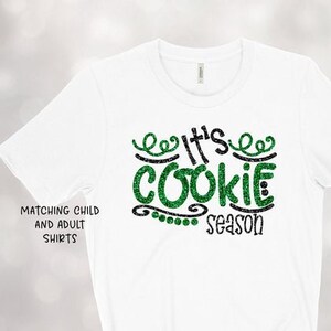 May include: White t-shirt with the text "It's Cookie Season" in green glitter lettering. The shirt also has the text "Matching Child and Adult Shirts" in black. The shirt is tied at the bottom.