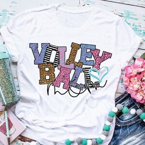 Pu&ograve; includere: Maglietta bianca con il testo "Volleyball Mom" in lettere colorate con un design a forma di cuore. Le lettere sono decorate con vari motivi, tra cui stampa leopardata, strisce e pois.