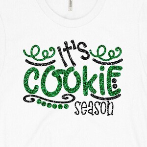 May include: White t-shirt with the text "It's COOKIE SEASON" in a glittery green and black design. The word "COOKIE" is in large, bold letters, and the text is surrounded by decorative swirls and dots. The shirt is made of a soft, comfortable material.