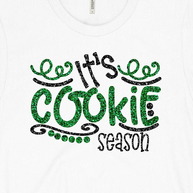 May include: White t-shirt with the text "It's COOKIE SEASON" in a glittery green and black design. The word "COOKIE" is in large, bold letters, and the text is surrounded by decorative swirls and dots. The shirt is made of a soft, comfortable material.