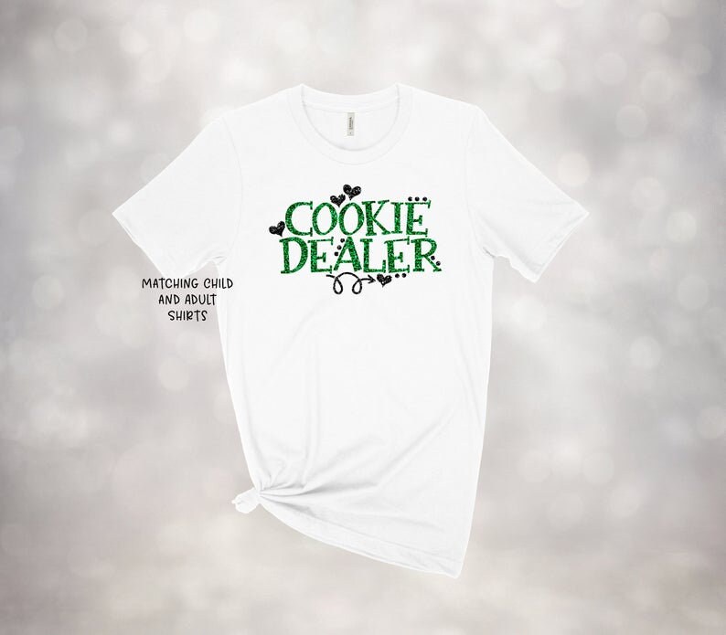 Puede incluir: Camiseta blanca con texto verde y negro que dice "Cookie Dealer" con corazones y una flecha. El texto es brillante. La camiseta tiene el texto "Matching Child and Adult Shirts" en la parte inferior izquierda.