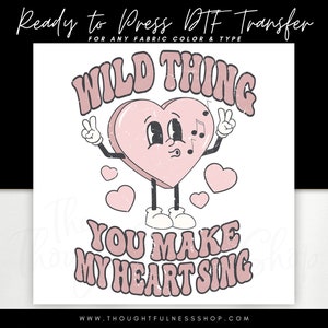 May include: A pink heart with a face and musical notes, with the text "WILD THING" above and "YOU MAKE MY HEART SING" below. The heart is wearing shoes and has arms outstretched with peace signs.