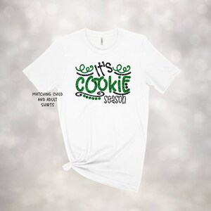 May include: White t-shirt with the text "It's Cookie Season" in green glitter and black lettering. The shirt has a crew neck and short sleeves. The words "Matching Child and Adult Shirts" are printed on the left side of the shirt.