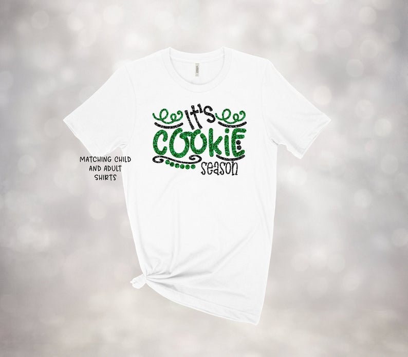 May include: White t-shirt with the text "It's Cookie Season" in green glitter lettering. The shirt also has the text "Matching Child and Adult Shirts" in black. The shirt is tied at the bottom.