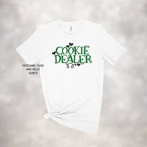 Puede incluir: Camiseta blanca con texto en purpurina verde que dice "Cookie Dealer" con corazones en purpurina negra. El texto tambi&eacute;n dice "Matching Child and Adult Shirts".
