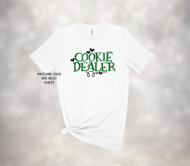 Puede incluir: Camiseta blanca con texto verde y negro que dice "Cookie Dealer" con corazones y una flecha. El texto es brillante. La camiseta tiene el texto "Matching Child and Adult Shirts" en la parte inferior izquierda.