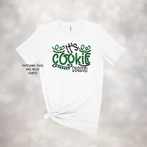 May include: White t-shirt with the text "It's Cookie Season" in green glitter and black lettering. The shirt also has the text "Matching Child and Adult Shirts" on the left side.