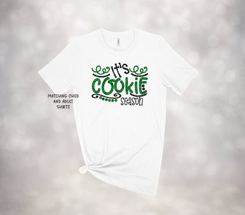 May include: White t-shirt with the text "It's Cookie Season" in green glitter and black lettering. The shirt also has the text "Matching Child and Adult Shirts" on the left side.