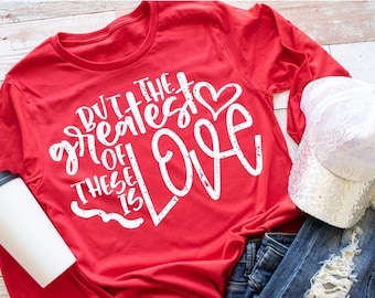 Puede incluir: Camiseta roja de manga larga con texto blanco que dice "Pero el más grande de estos es el amor" con un gráfico de corazón blanco.
