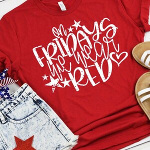 May include: Red t-shirt with white text that says "On Fridays we wear red" with stars and a heart. The shirt is laying on a wooden surface with a pair of sandals and a pair of denim shorts.