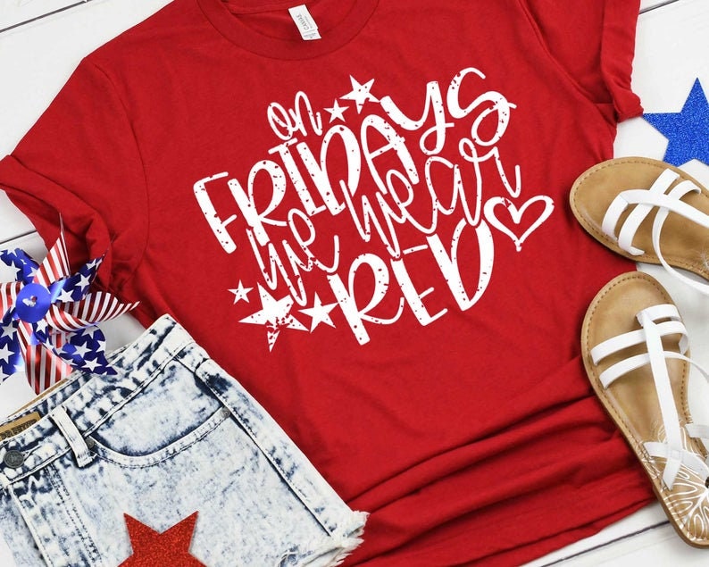 May include: Red t-shirt with white text that says "On Fridays we wear red" with stars and a heart. The shirt is laying on a wooden surface with a pair of sandals and a pair of denim shorts.