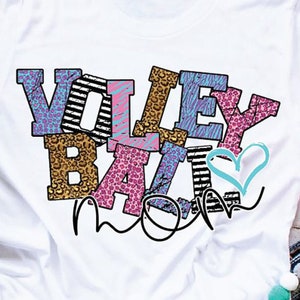 Pu&ograve; includere: Maglietta bianca con le parole "Volley Ball Mom" in lettere colorate con vari motivi. Le lettere sono in un design a strati e sovrapposti, con una forma di cuore al centro.