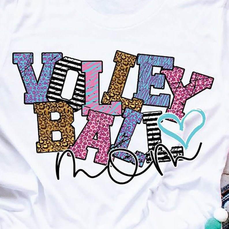Pu&ograve; includere: Maglietta bianca con le parole "Volley Ball Mom" in lettere colorate con vari motivi. Le lettere sono in un design a strati e sovrapposti, con una forma di cuore al centro.