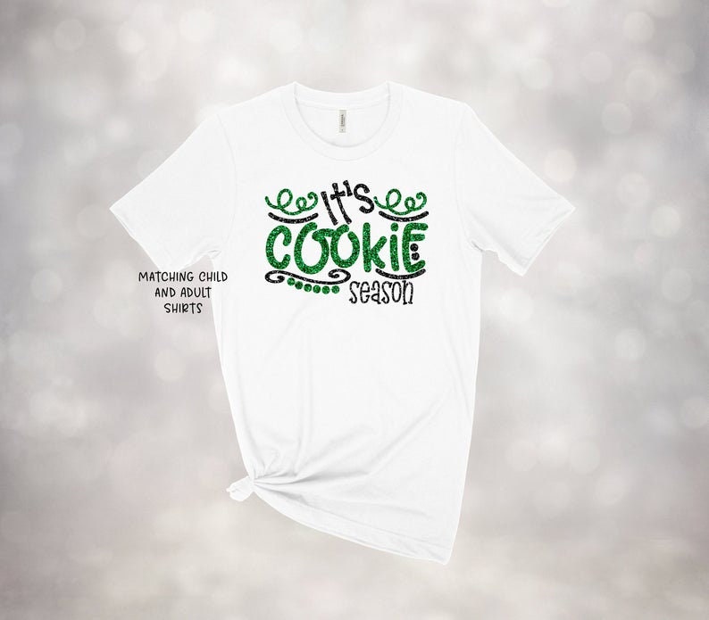 May include: White t-shirt with the text "It's Cookie Season" in green glitter and black lettering. The shirt also has the text "Matching Child and Adult Shirts" on the left side. The shirt is tied at the bottom.