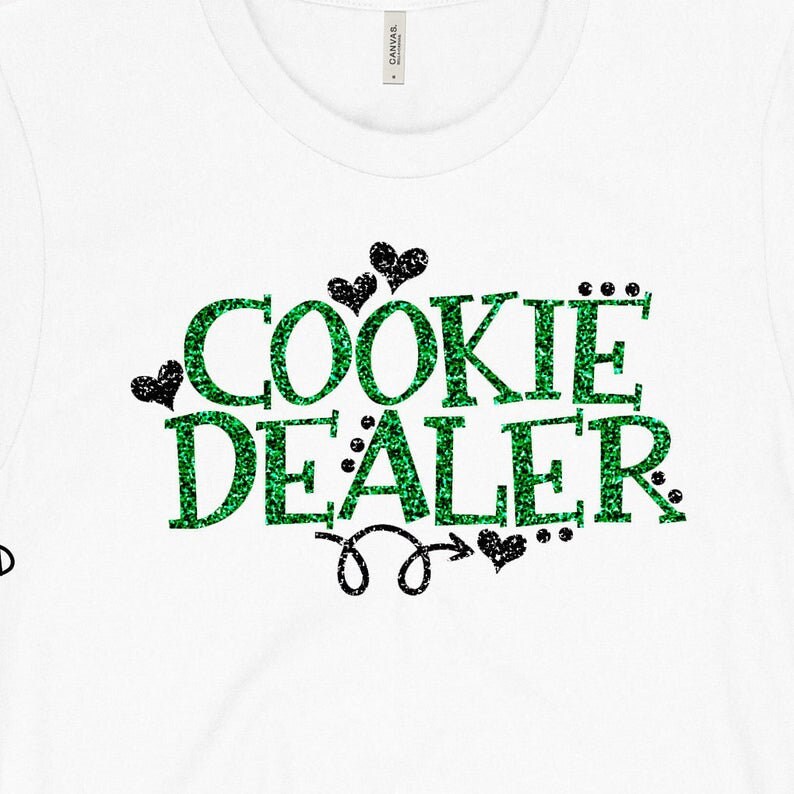 Puede incluir: Camiseta blanca con texto en purpurina verde que dice "COOKIE DEALER" con corazones en purpurina negra y una flecha.