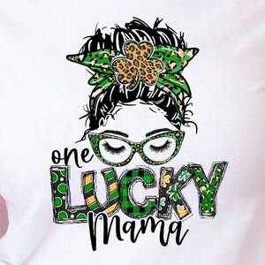 May include: A white t-shirt with a green, black, and gold graphic design. The graphic features a woman with a messy bun, glasses, and a shamrock headband. The text "one lucky mama" is written in a green, black, and gold font.