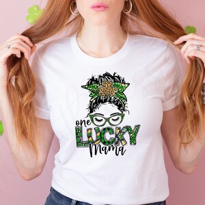 May include: White t-shirt with a green, gold, and black graphic of a woman with a shamrock headband and the text "one lucky mama".